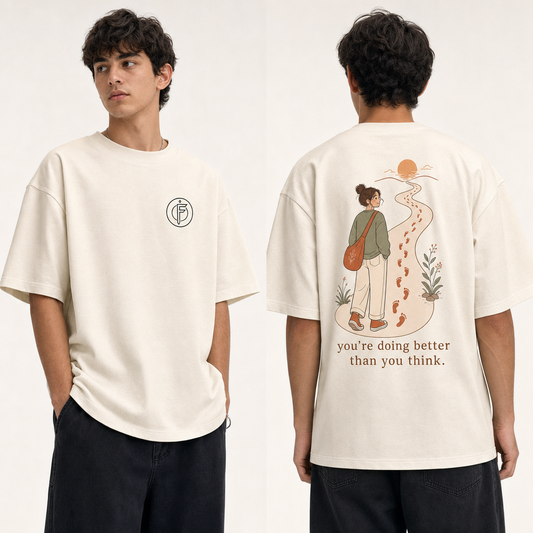 Editorial studio photo of a model wearing an oversized off-white heavyweight t-shirt with a graphic reading YOU ARE DOING BETTER THAN YOU THINK, relaxed streetwear fit, clean studio background.